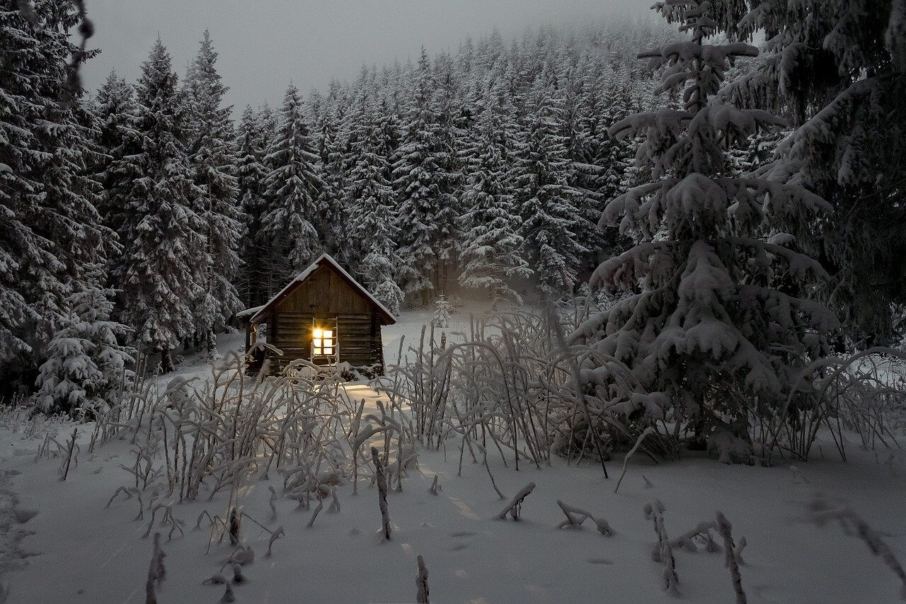 domek na pokrytej śniegiem polanie. W okienku pali się światło. W tle zaśnieżone sosny.